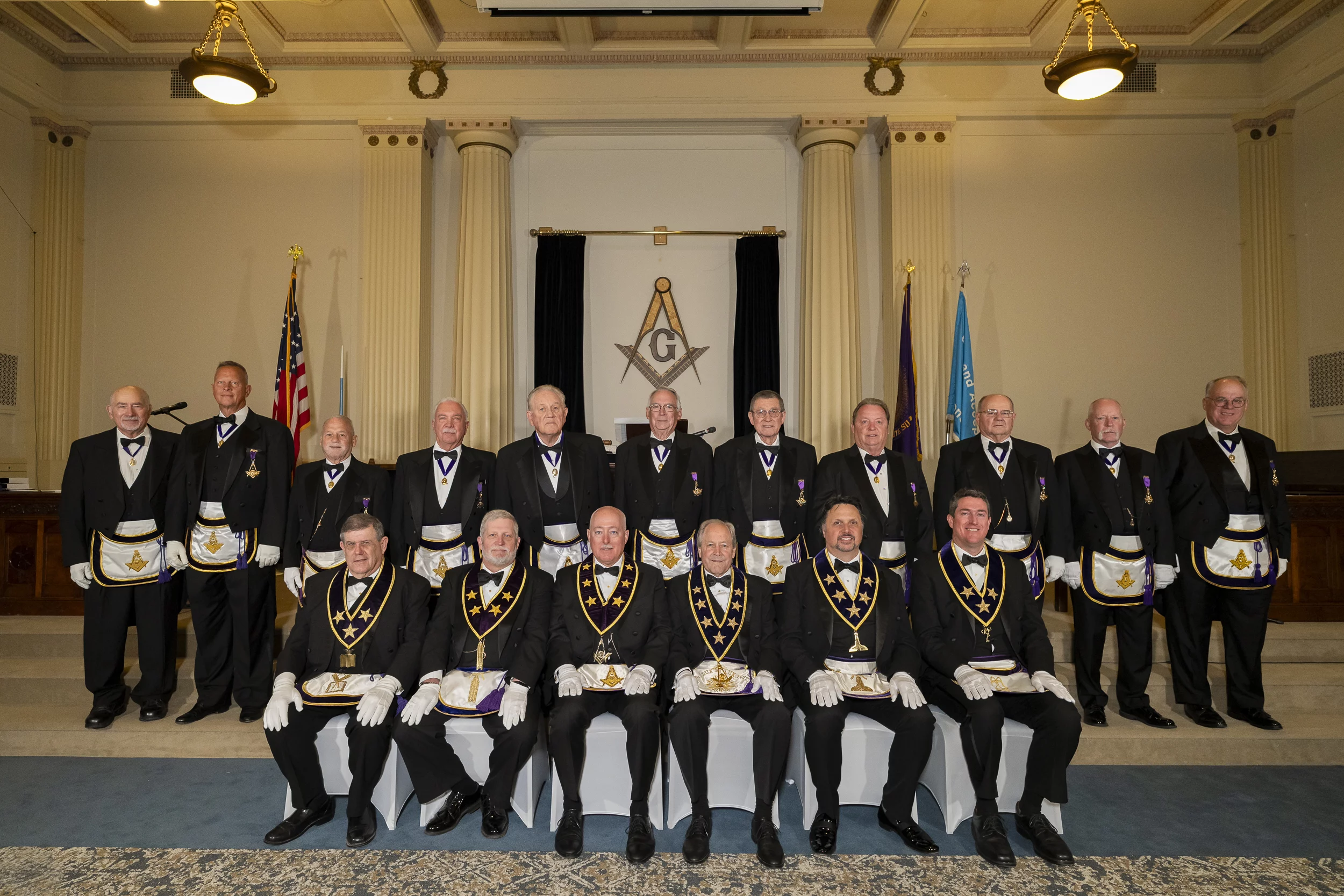 Group of Masons sitting and standing together with Grand Lodge officers seated in front.
