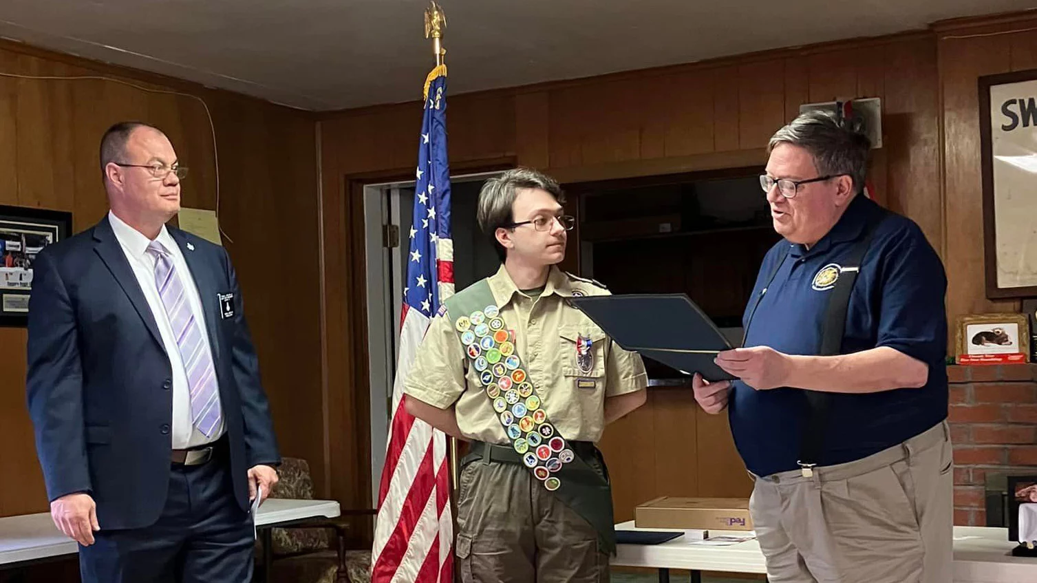 Man presenting an award to teen dressed in boy scout uniform while his father stands behind him.