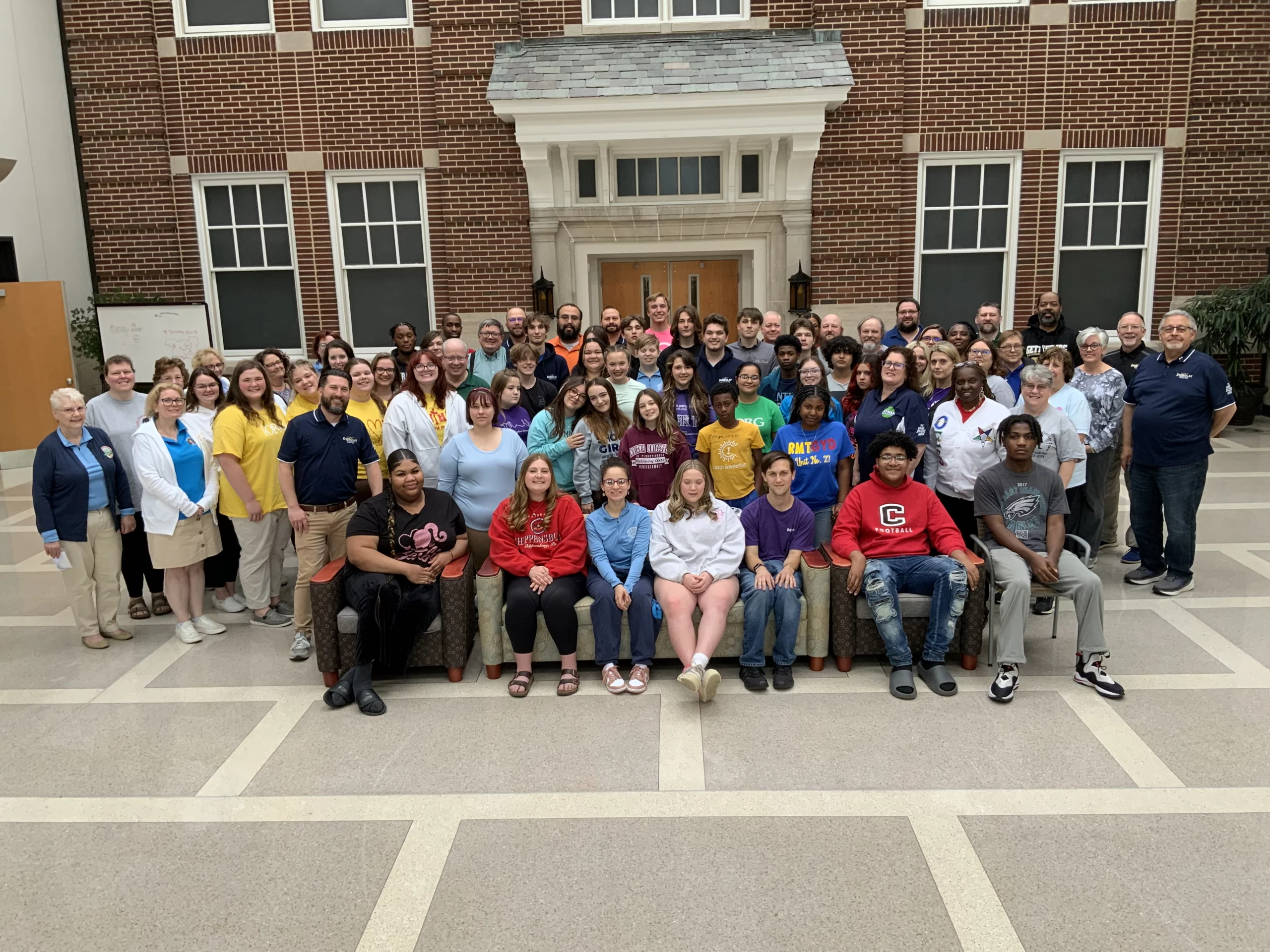 Large group of teens, children, and adults in front of brick building.