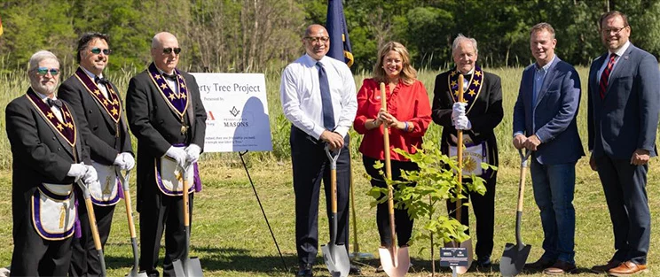 Group of people dressed in mix of formal and Masonic dress standing behind newly planted tree.