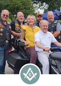 Grand Master Larry Derr and his wife sitting on motorcycle surrounded by men dressed in motorcycle gear with Square and Compass icon.