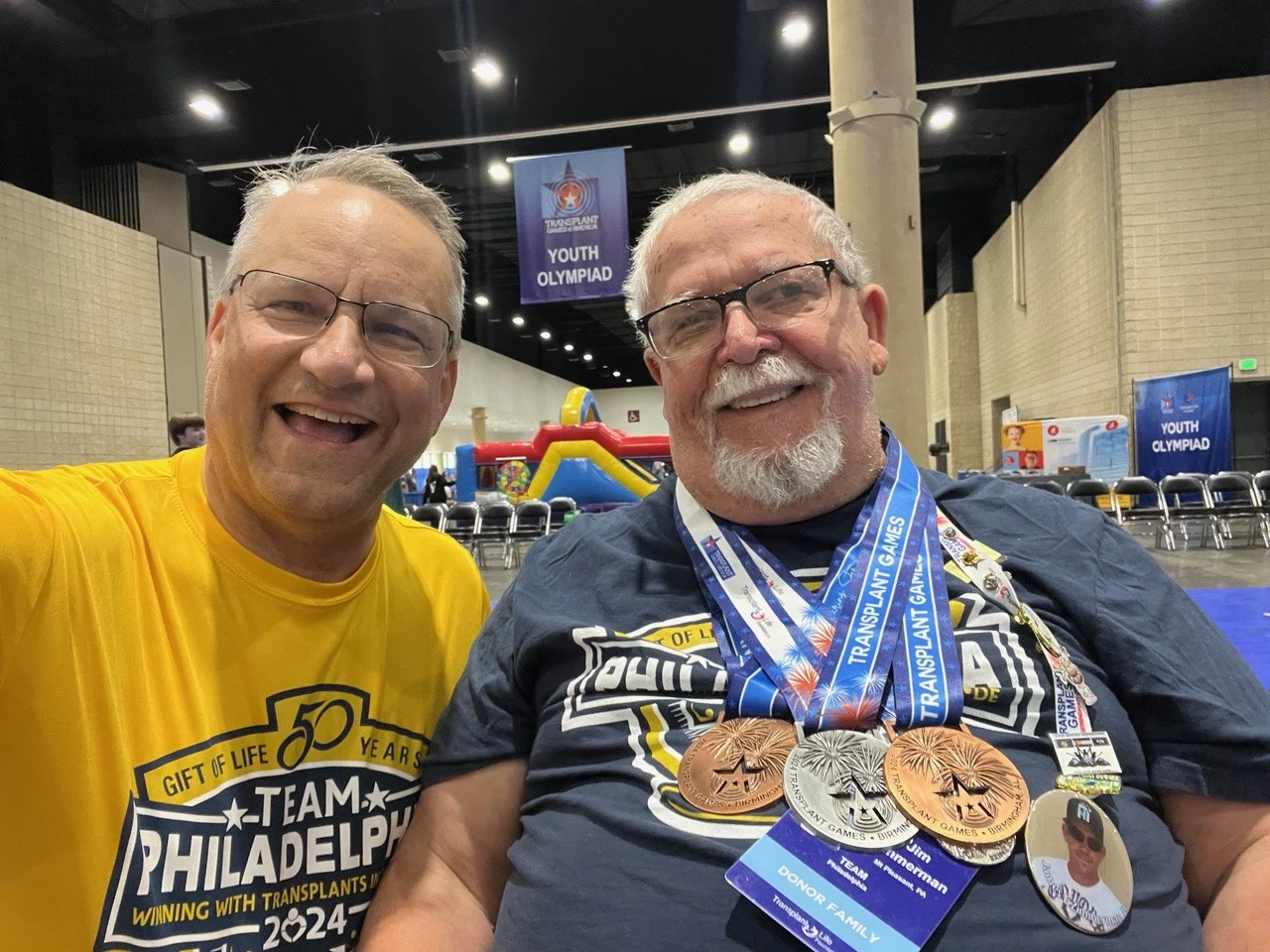bill and james 1 Two smiling men with Team Philadelphia shirts on and metals from sport events.