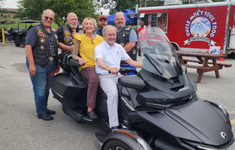 20240803_092120 Grand Master Larry Derr with his wife sitting on 3-wheeled motorcycle with men dressed in motorcycle riding gear behind them.