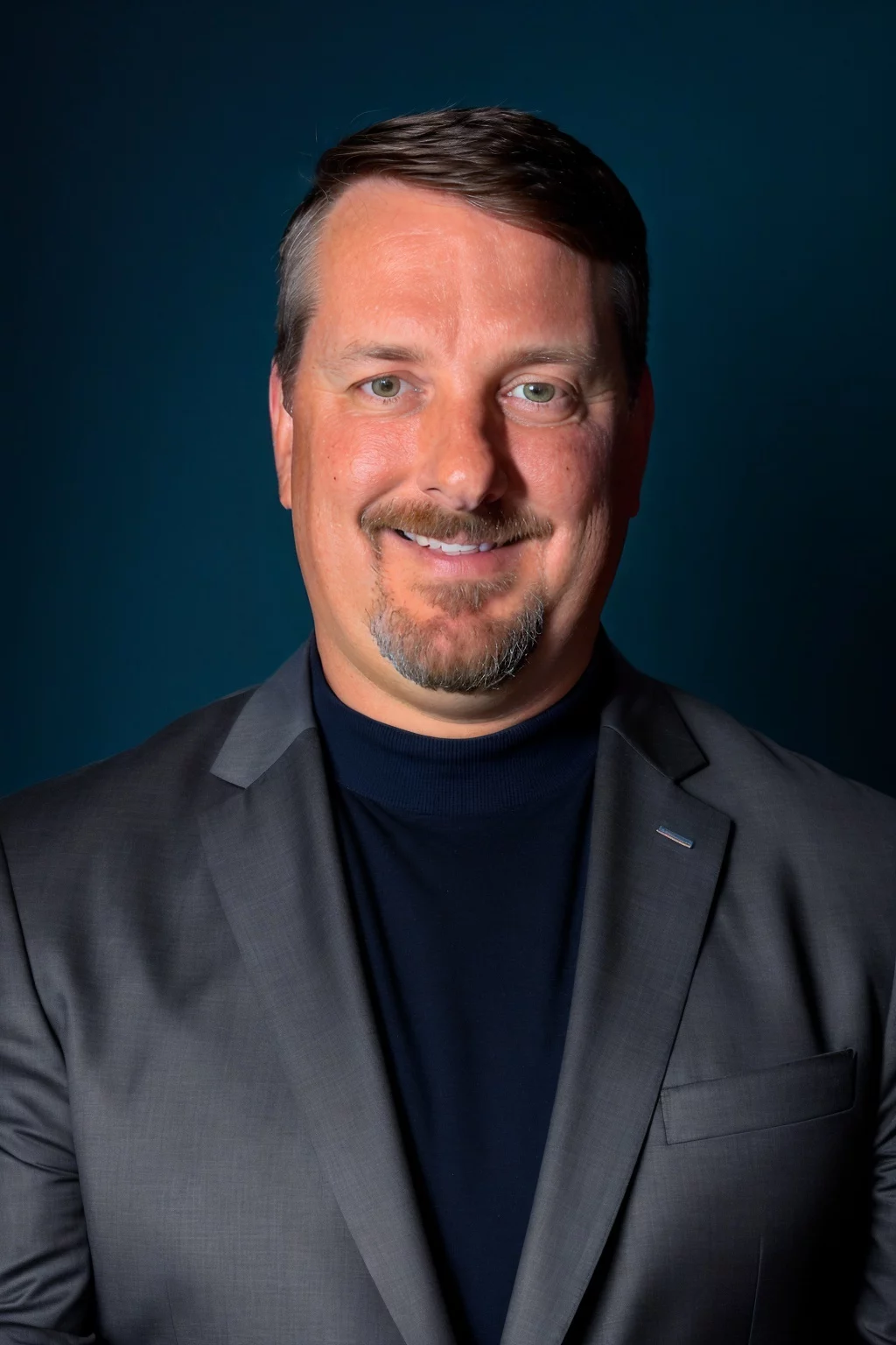 Greg Tadda Headshot of smiling man with short brown hair and goatee wearing a blazer.