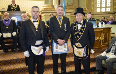Small group of men standing in ornate hall with seated crowd behind them.