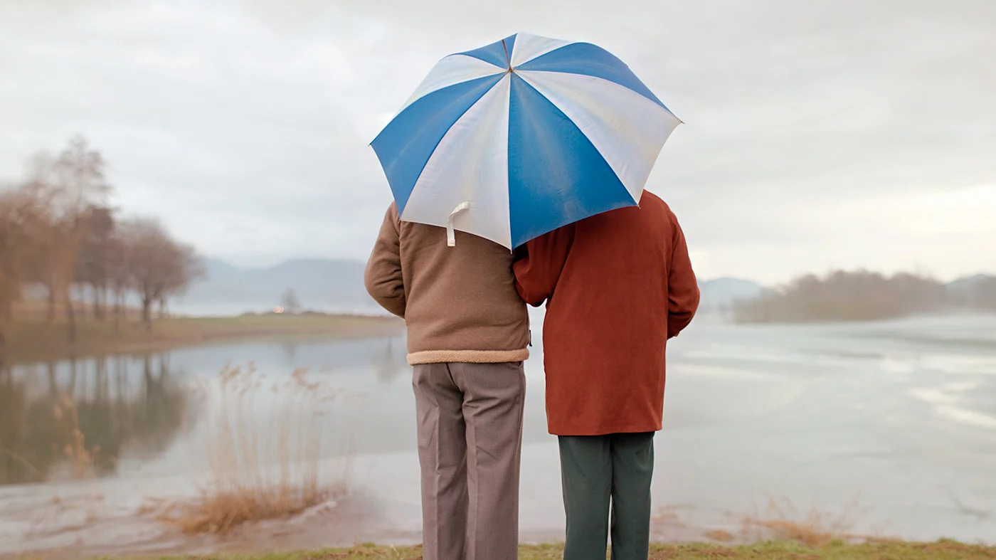 Couple standing on edge of lake with umbrella over their heads.