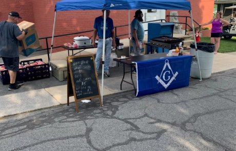 45-2 Men standing under outdoor tent with BBQ on a grill.