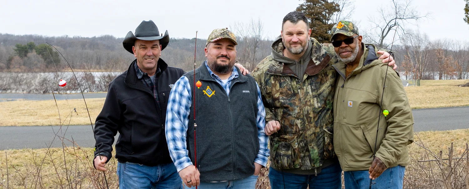 Group of smiling men with fishing equipment standing together outdoors.