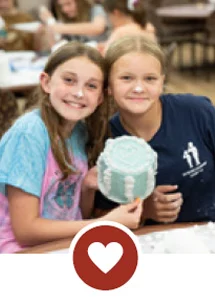 Smiling girls holding up blue craft project with heart icon.