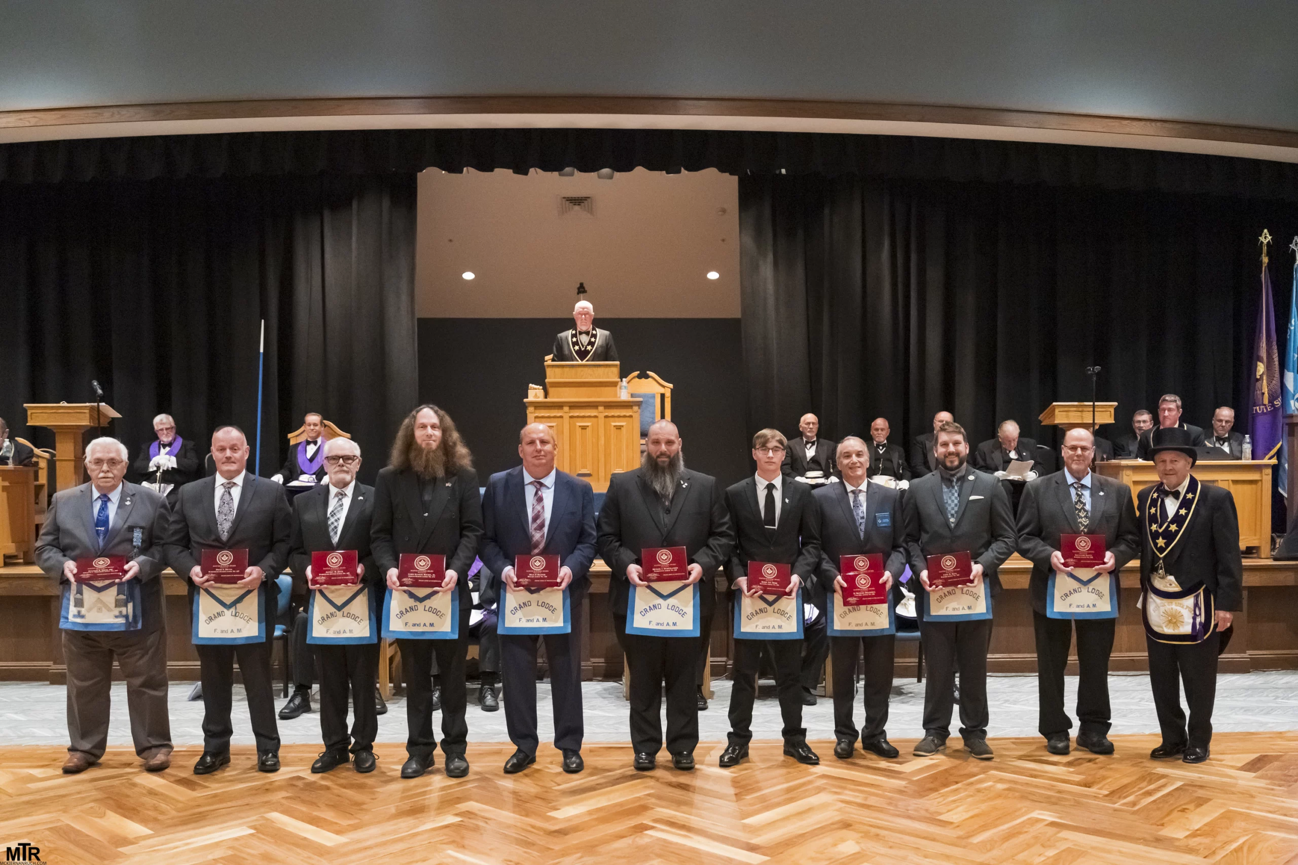 Group of Masons standing together with red plaques, Grand Master Larry Derr is standing with them to the side..