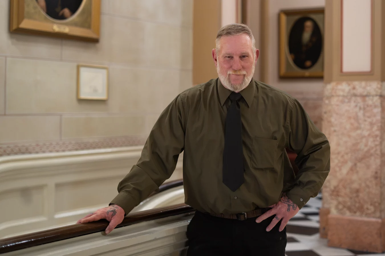Smiling man in green shirt and black tie standing in hall with portraits behind him.