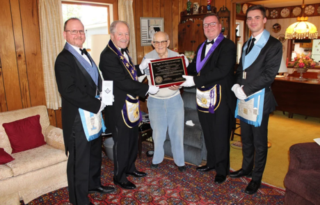 Group of men in masonic dress presenting man with black and wood plaque in home.