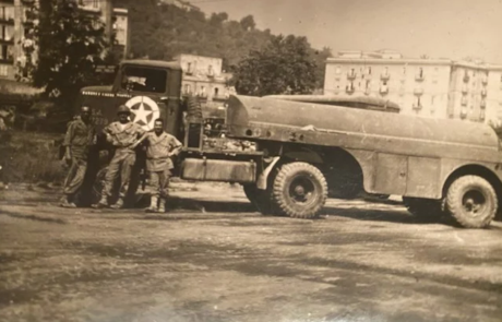 Aged photo of men in military uniform standing in front of large truck.
