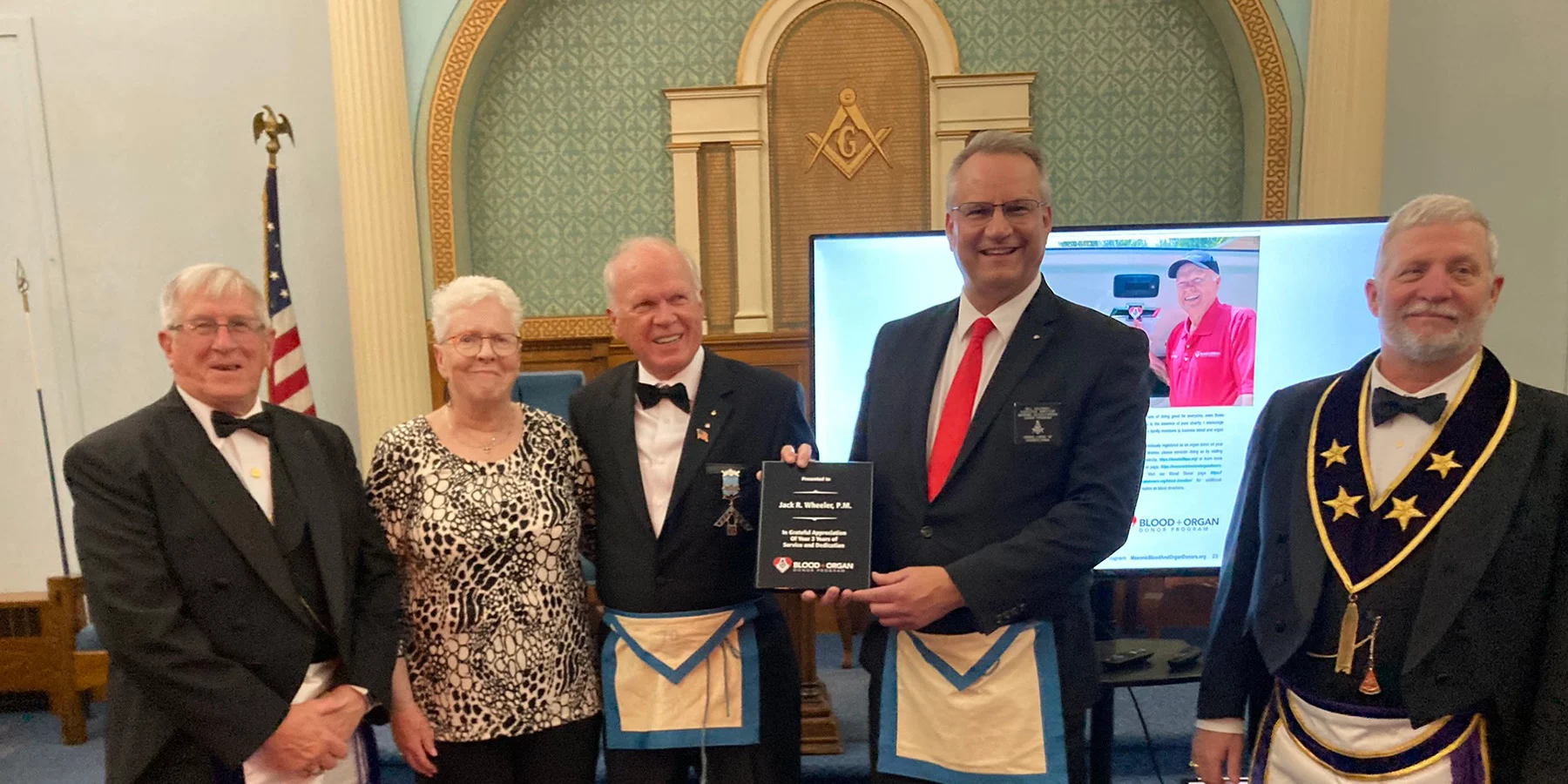 Group of men in masonic dress and woman smiling, one man holding up award for Jack Wheeler.
