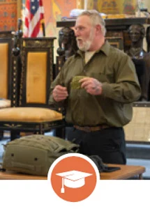 Man lecturing in ornate lodge hall with graduation cap icon.