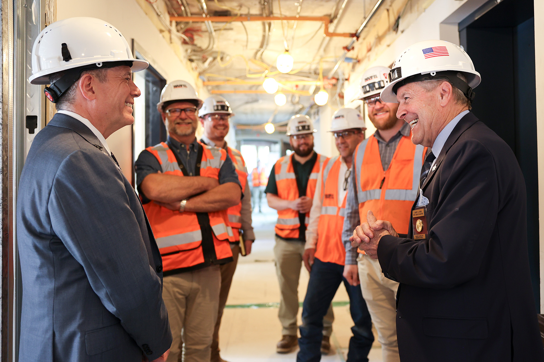 Grand Master Larry Derr smiling while talking to man in a suit and men in construction vests in hallway