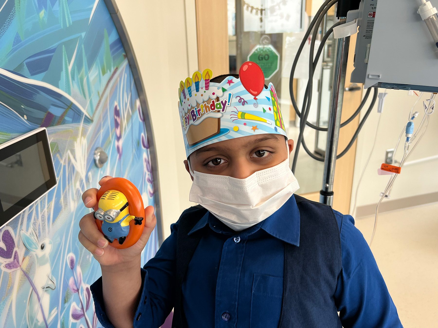 Boy in mask with birthday hat holding up toy minion