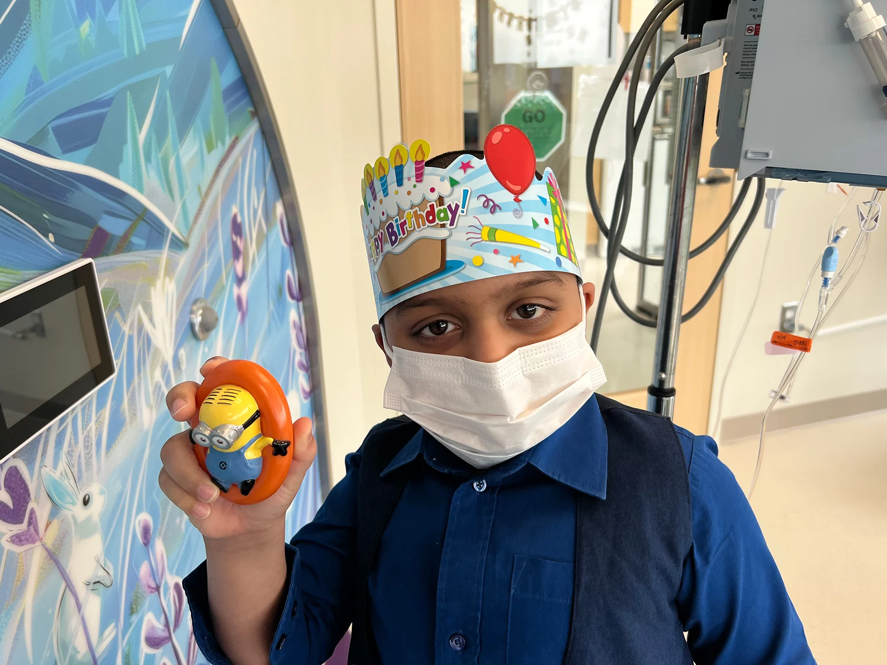 Boy in mask with birthday hat holding up toy minion.