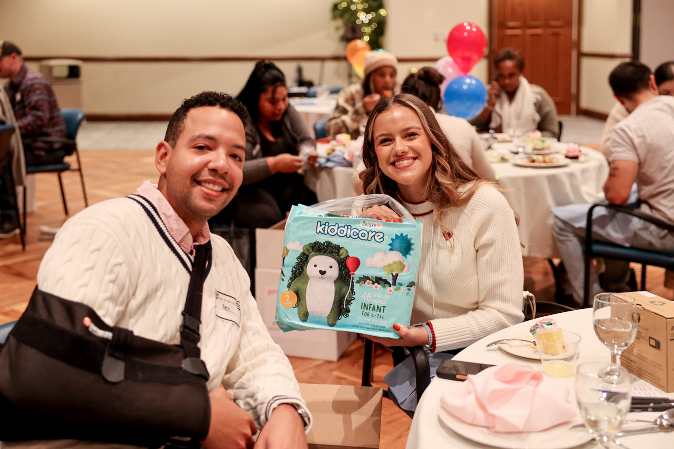 1W8A4980 Smiling couple seated at table while holding up package of baby supplies.