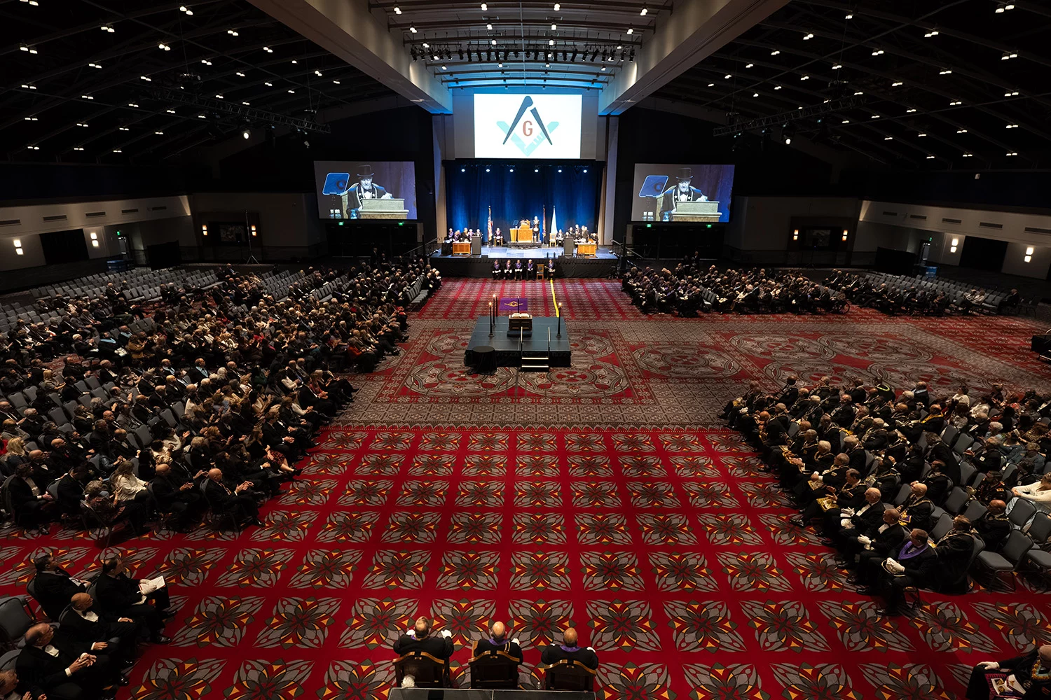 Large lodge room with Masons seated at either side and man presenting on stage,