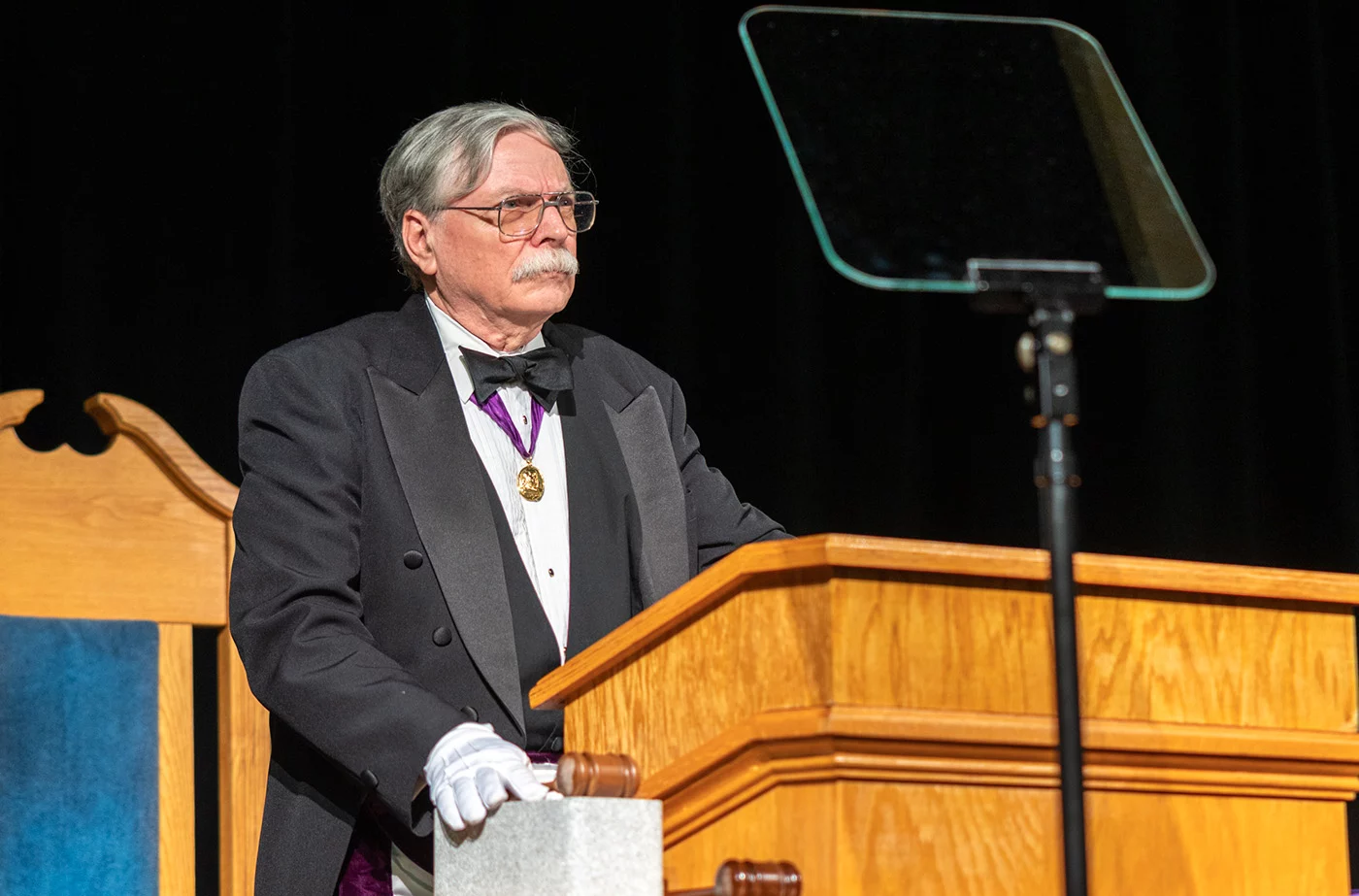 Man with grey hair, glasses and mustache in Masonic attire standing at a podium.