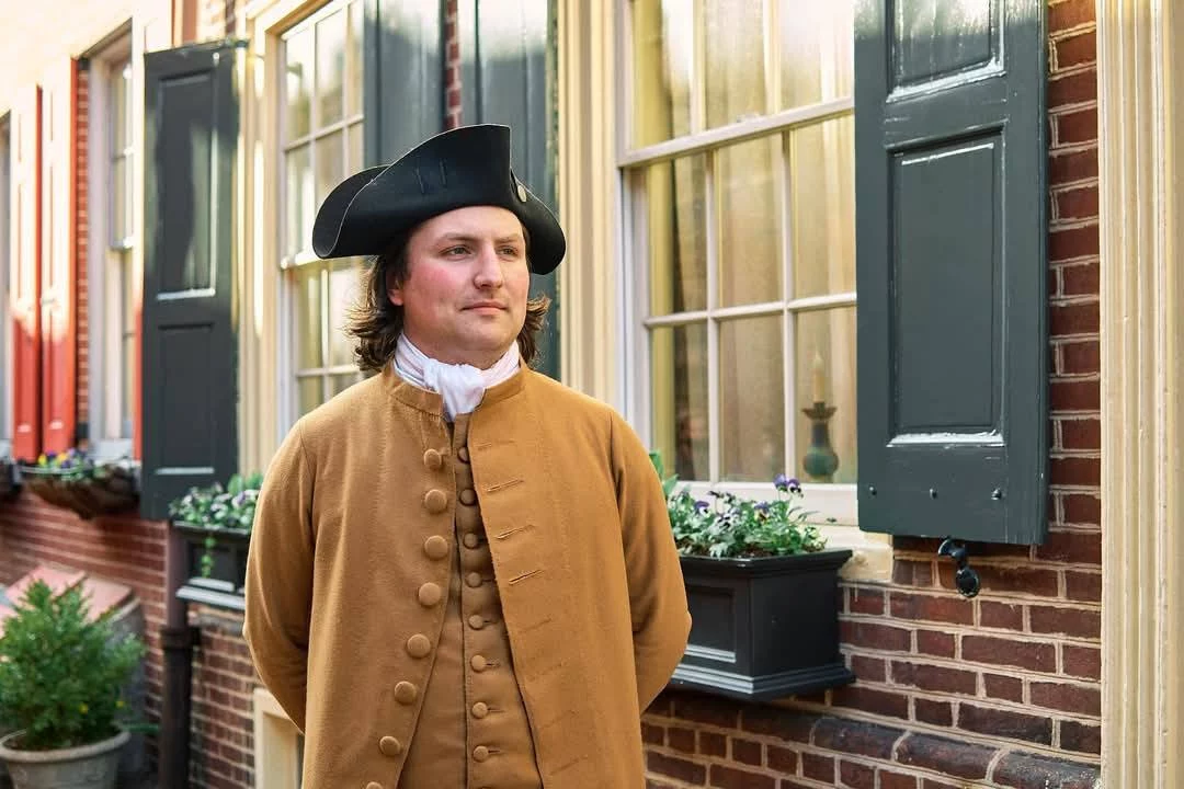 Man in Colonial era clothing standing in front of brick building.