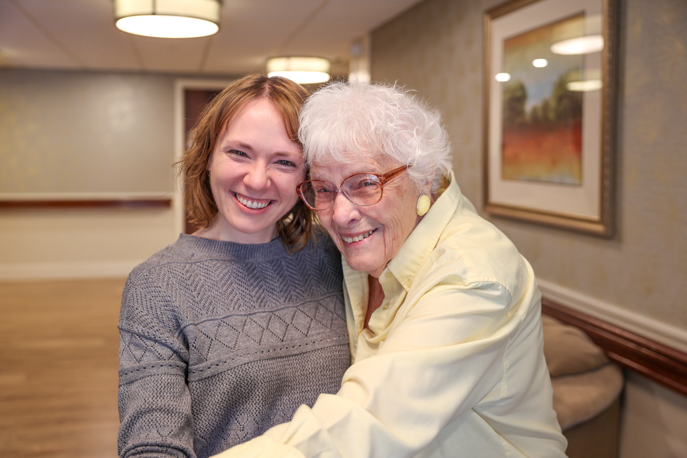 Smiling senior woman in glasses hugging young woman while standing in a hallway together.