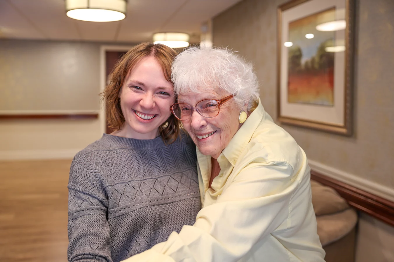 Smiling senior woman in glasses hugging young woman while standing in a hallway together.