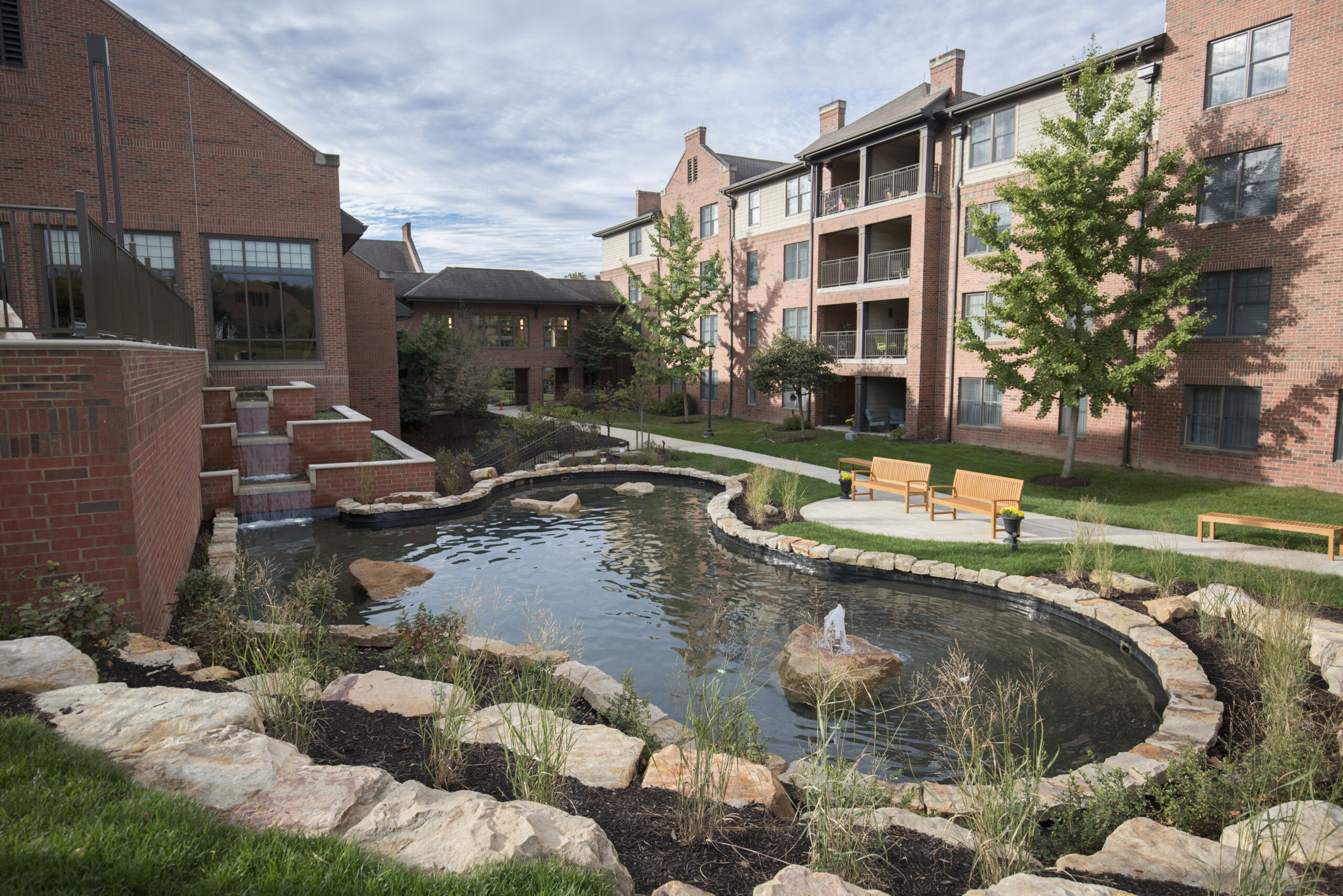 Quaint pond with small fountain surrounded by stones and benches next to large brick building.