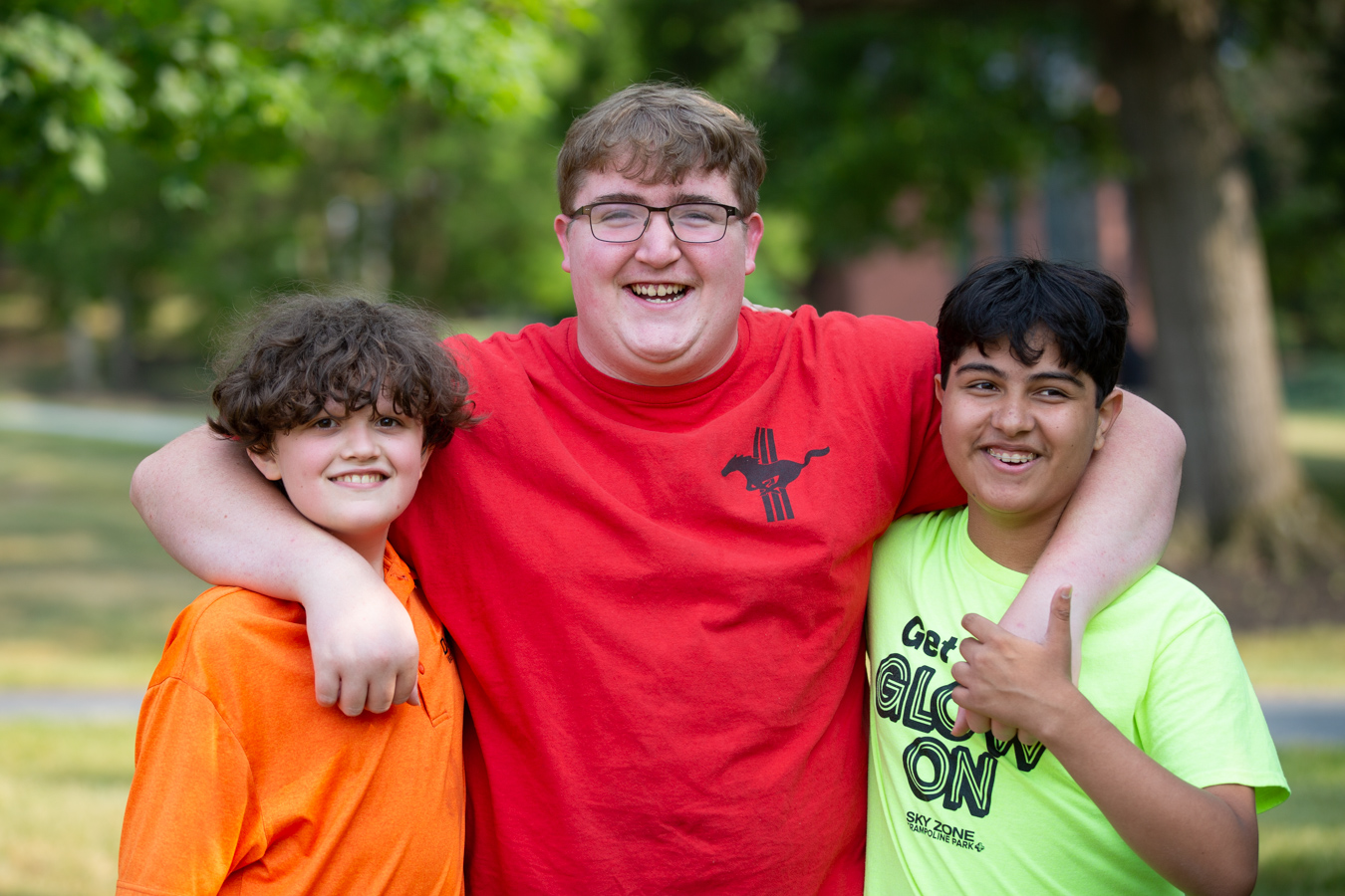 Young man with his arms around two younger boys, all are smiling while standing outdoors.