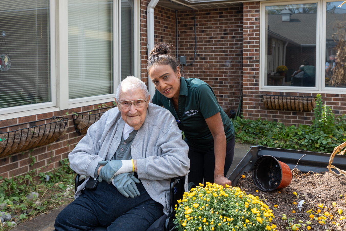 9I4A7383 Smiling senior man seated in a wheelchair wearing gardening gloves. A smiling woman in a green polo is standing behind him. A raised garden bed is beside them.