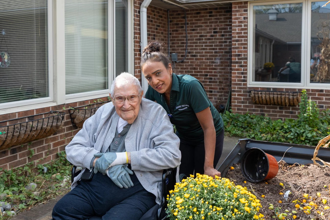Smiling senior man seated in a wheelchair wearing gardening gloves. A smiling woman in a green polo is standing behind him. A raised garden bed is beside them.
