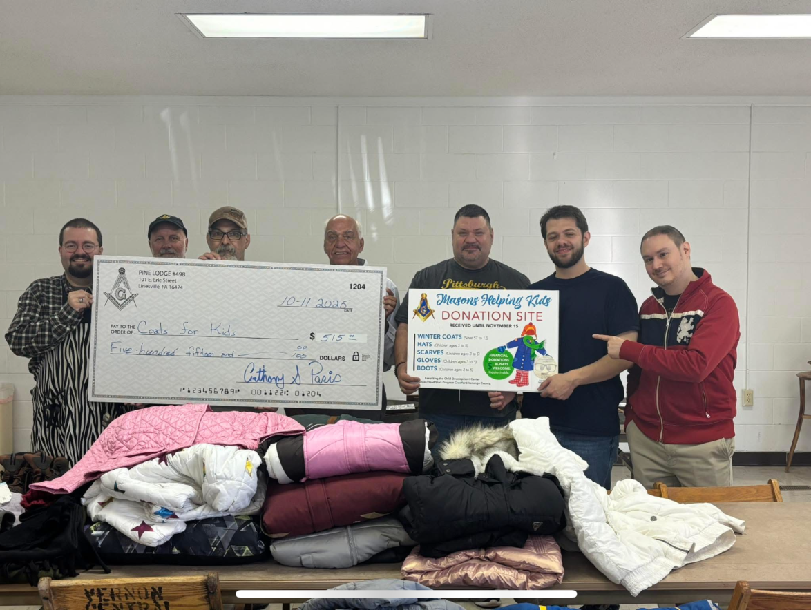 9bcca84f-4c57-415b-bada-1637319c9997 (1) Group of smiling men holding up a giant check and sign reading, "Masons Helping Kids," while standing in front of pile of coats.