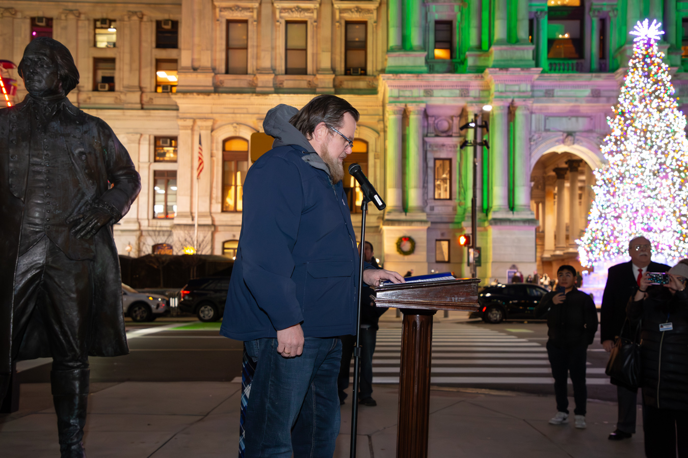 Michael McKee, a man with brown hair, wearing glasses and a winter coat while speaking at a podium outside. A large, illuminated Christmas tree and Philadelphia's City Hall are in the background.