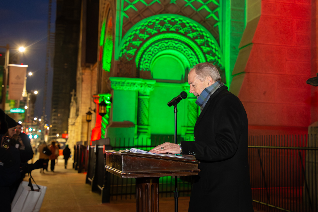 Grand Master Larry Derr, a man with gray hair, wearing a blue scarf and black jacket, speaking at a podium outdoors. The illuminated Masonic Temple, Library and Museum walls and the sidewalks of a city street are in the background.