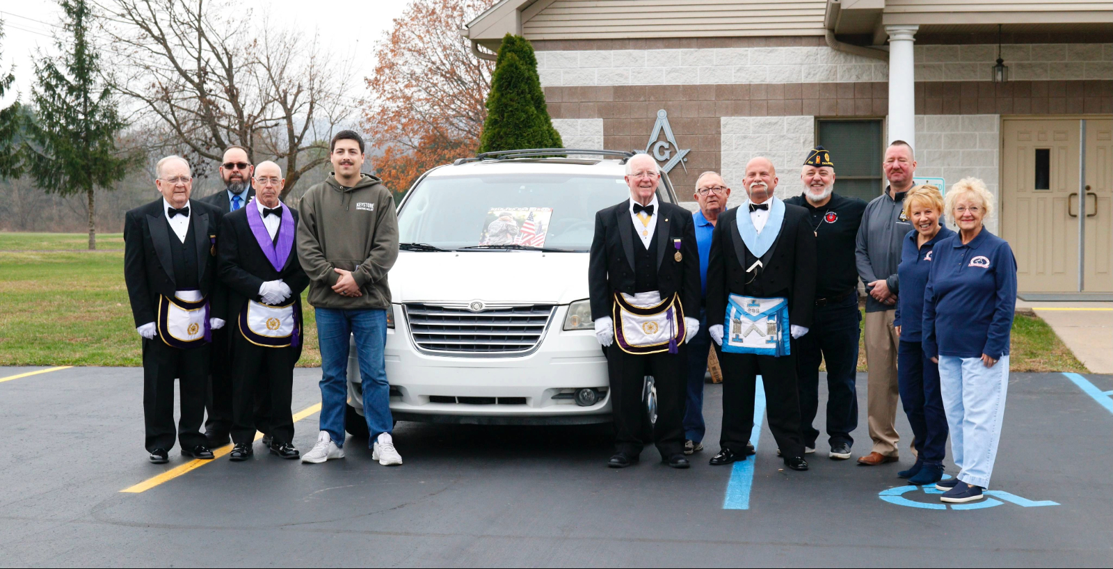 Smiling group, some of whom are in Masonic dress standing outside by white van.