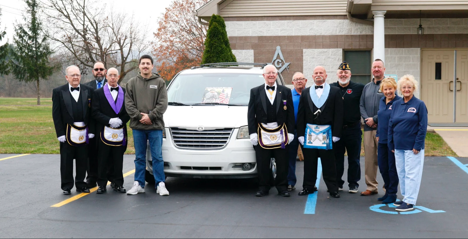 Smiling group, some of whom are in Masonic dress standing outside by white van.