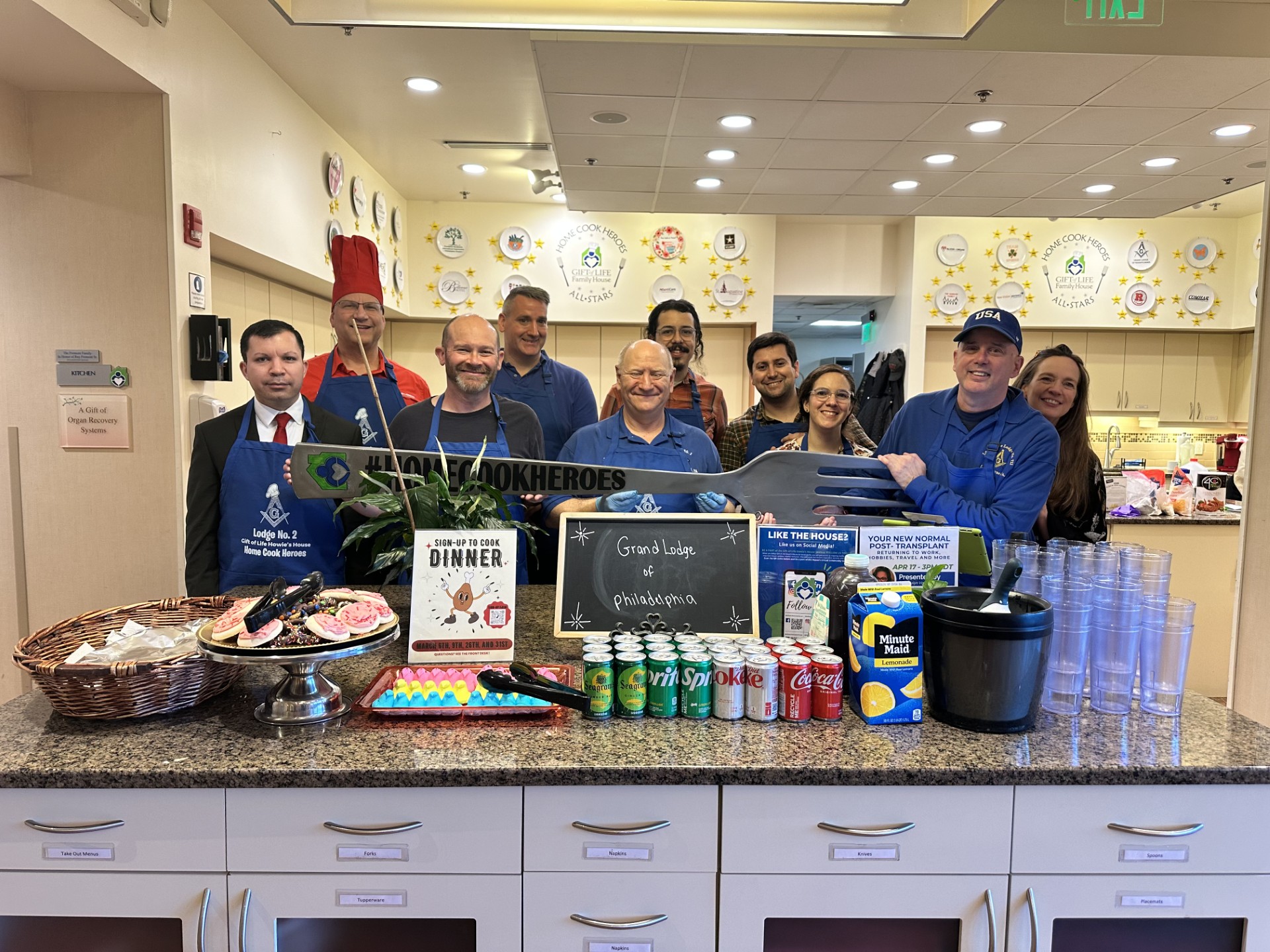 Group of volunteers holding a fork standing in a kitchen with drinks and snacks on counter in front of them.