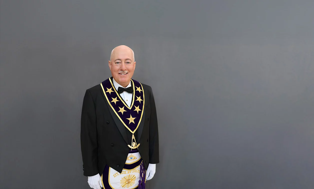 Portrait of Grand Master Robert Brink, a mature man with a mustache dressed in Masonic regalia, smiling and standing against a grey background.