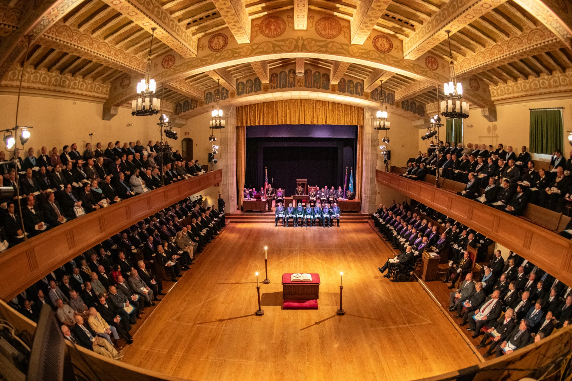 Large, ornate room with Masons seated on multi-level benches on either side. A small group of men is seated by the stage in the center.