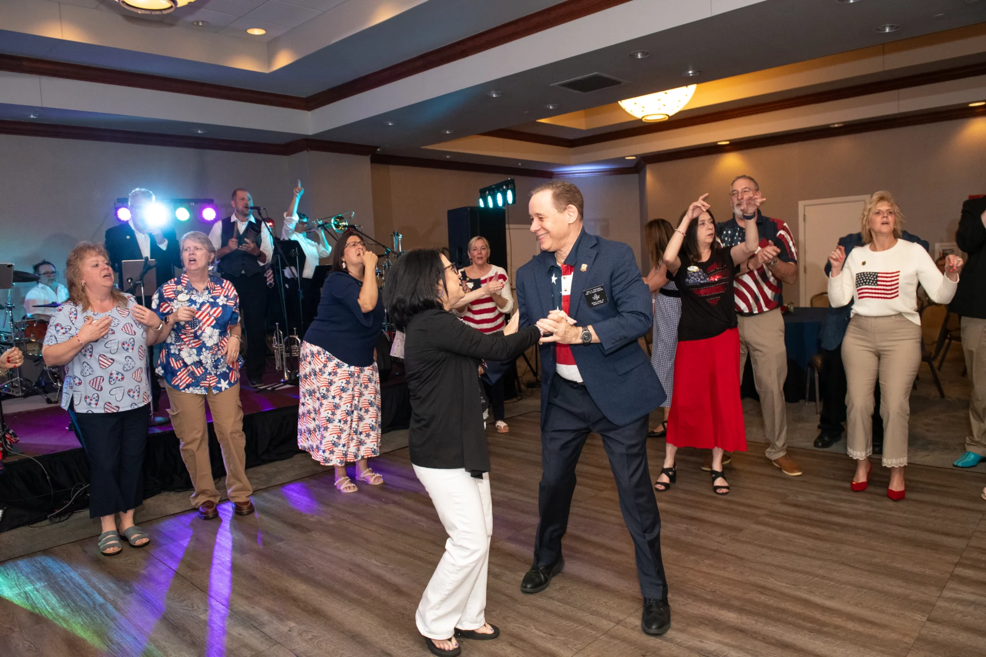 Smiling man and woman dancing together in a ballroom, a DJ and other people dancing are in the background.