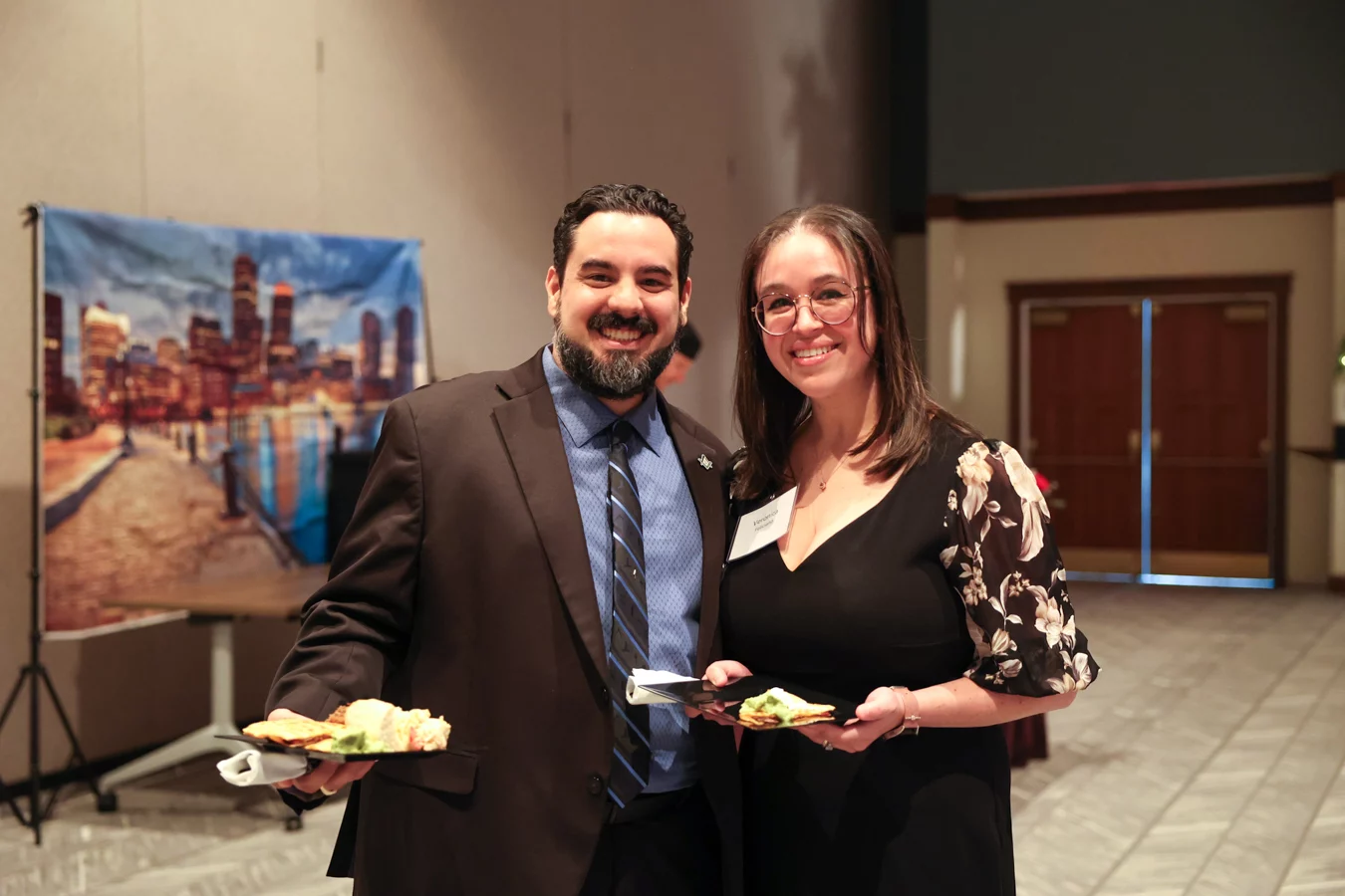 1W8A7178 Smiling man and woman in formal attire holding plates of food.
