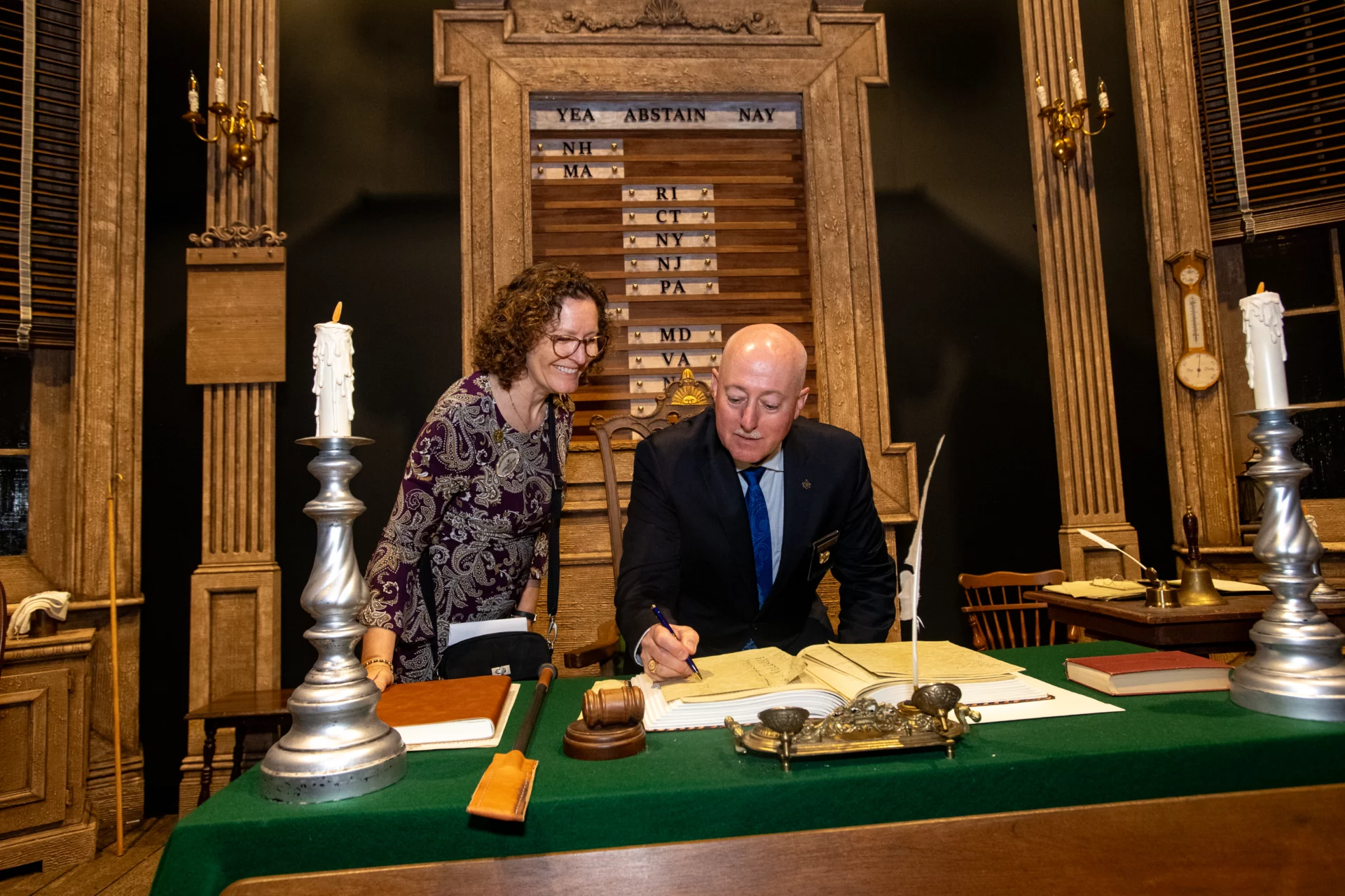 Grand Master Robert Brink signing document at an aged desk, with his smiling wife standing beside him.