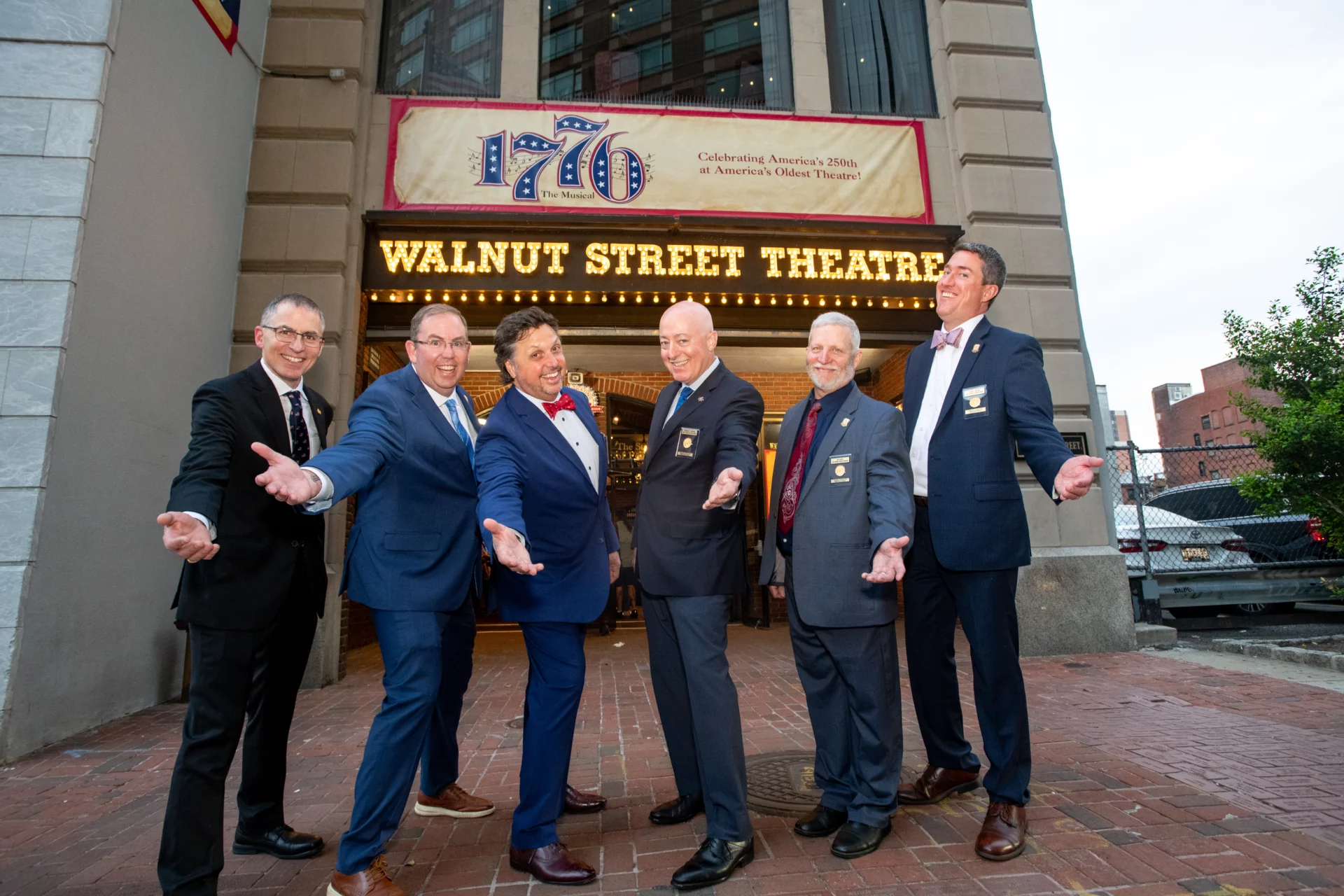 Grand Lodge Officers dressed in formal attire with their arms out in front of Walnut Street Theatre's 1776 Sign.