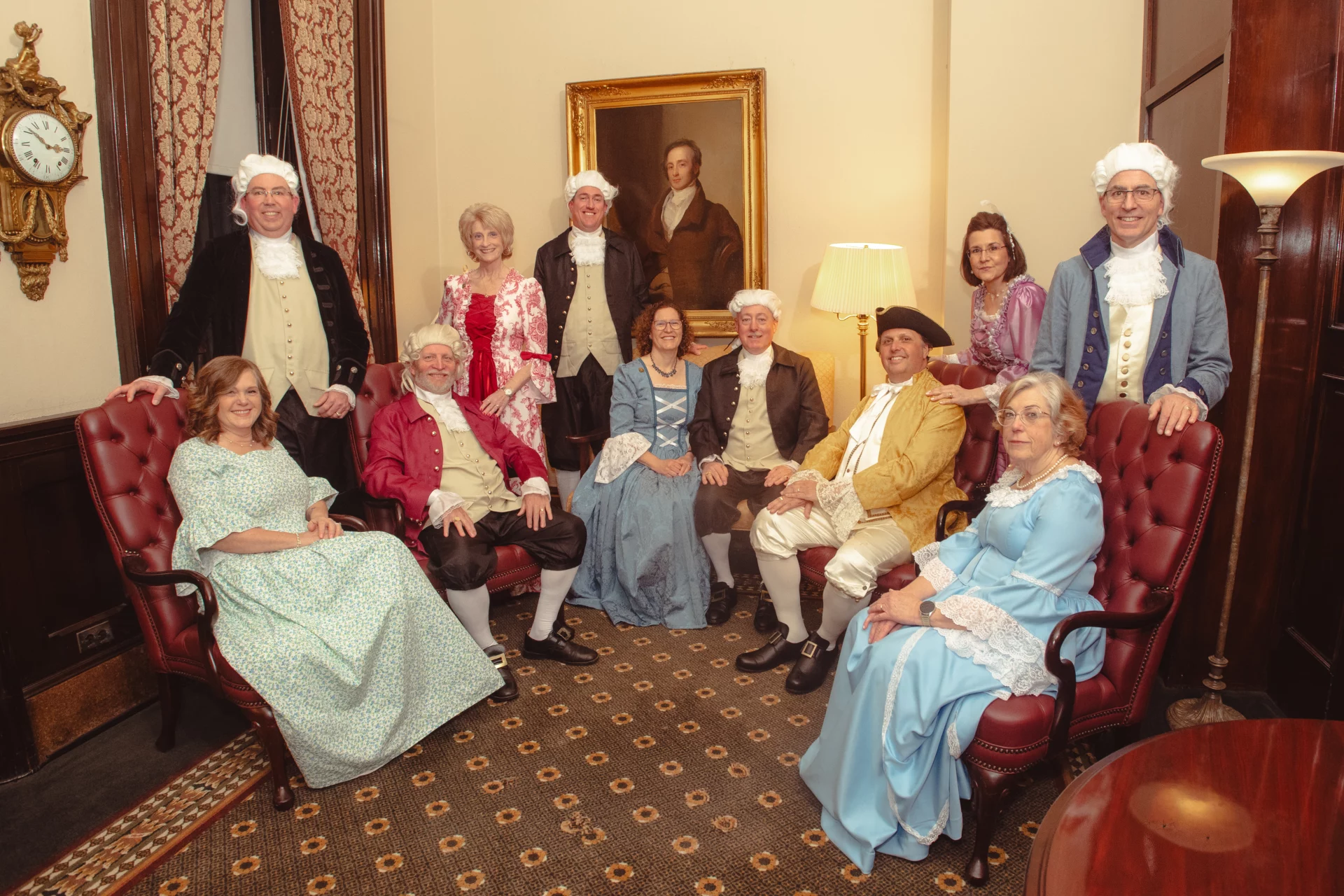 Group of men and women standing together in an ornated room, dressed in colonial outfits.