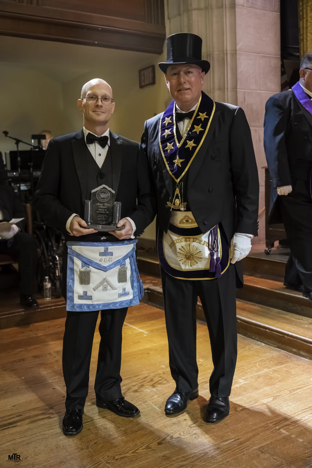 Grand Master Robert Brink standing next to a man holding up an award, both are dressed in Masonic attire.