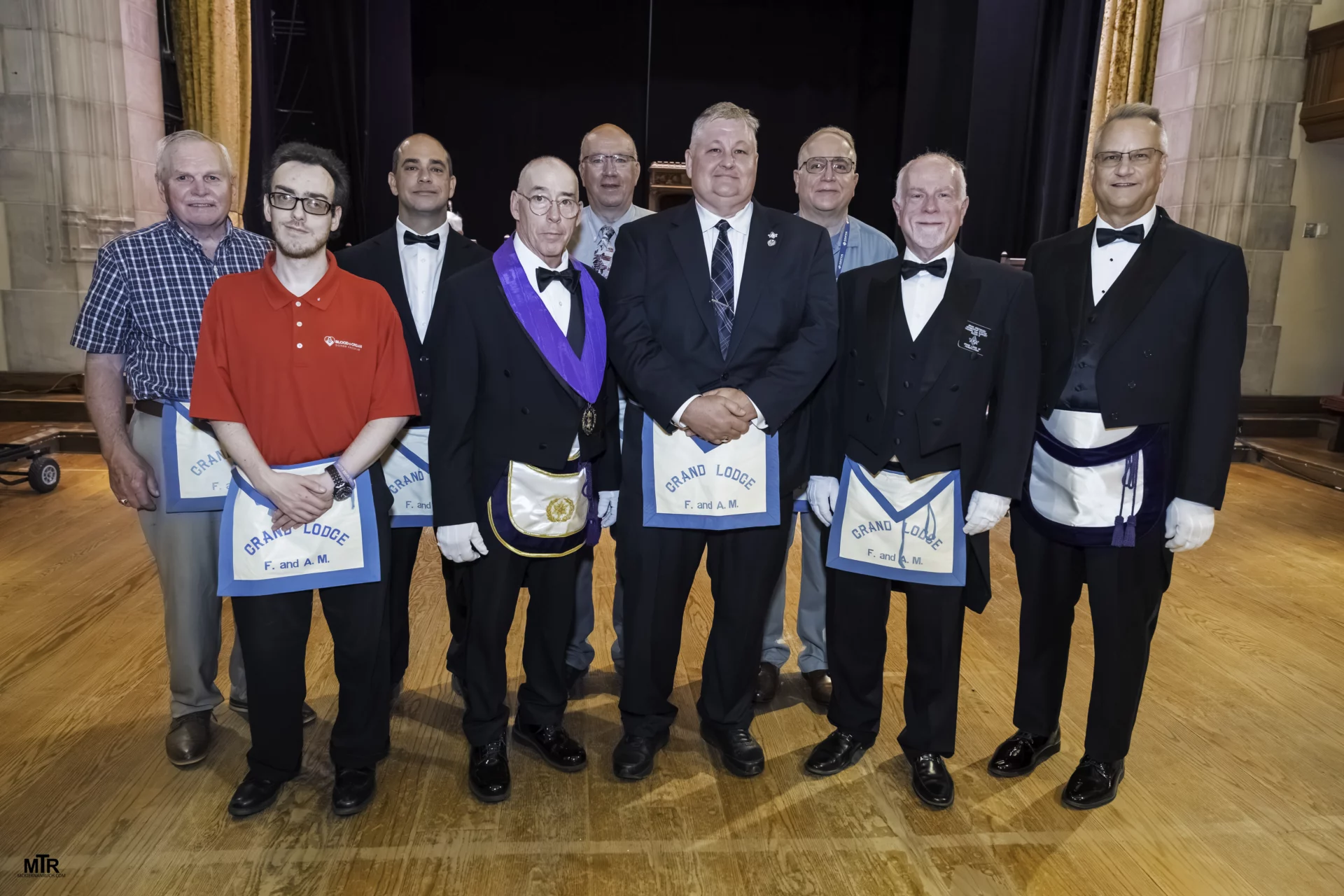 Smiling group of men standing in a large room together, all are wearing Masonic Aprons.