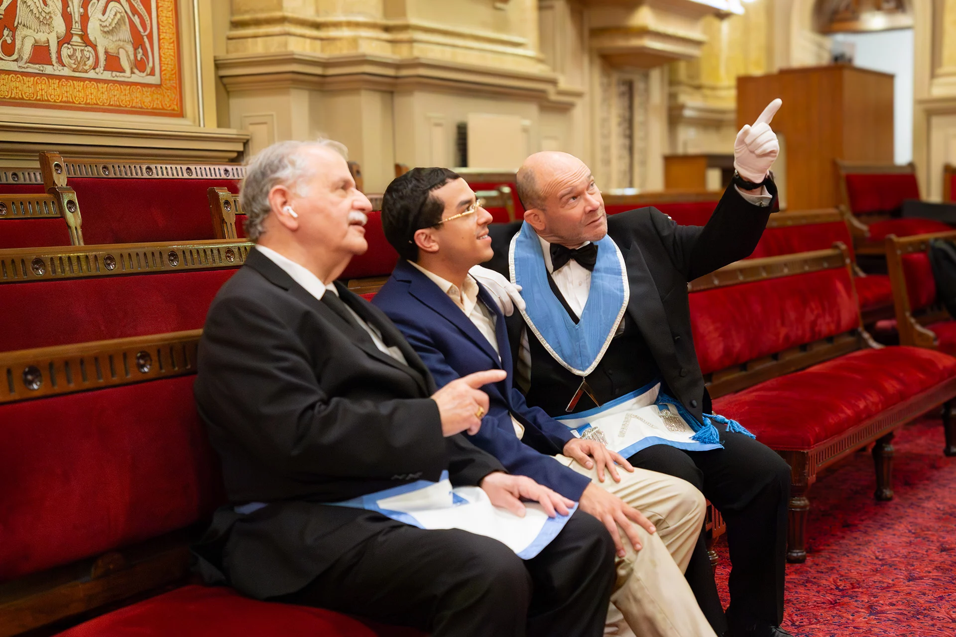 Three smiling men seated on a bench together in a lodge room, one man is pointing out details to a younger member.