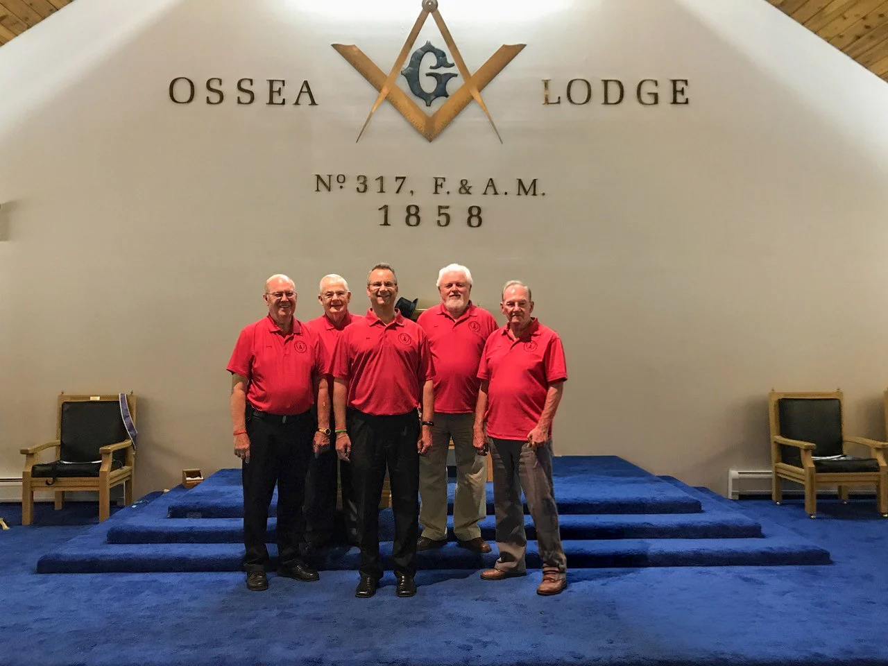 Smiling group of men standing in front of Ossea Lodge sign while standing indoors.