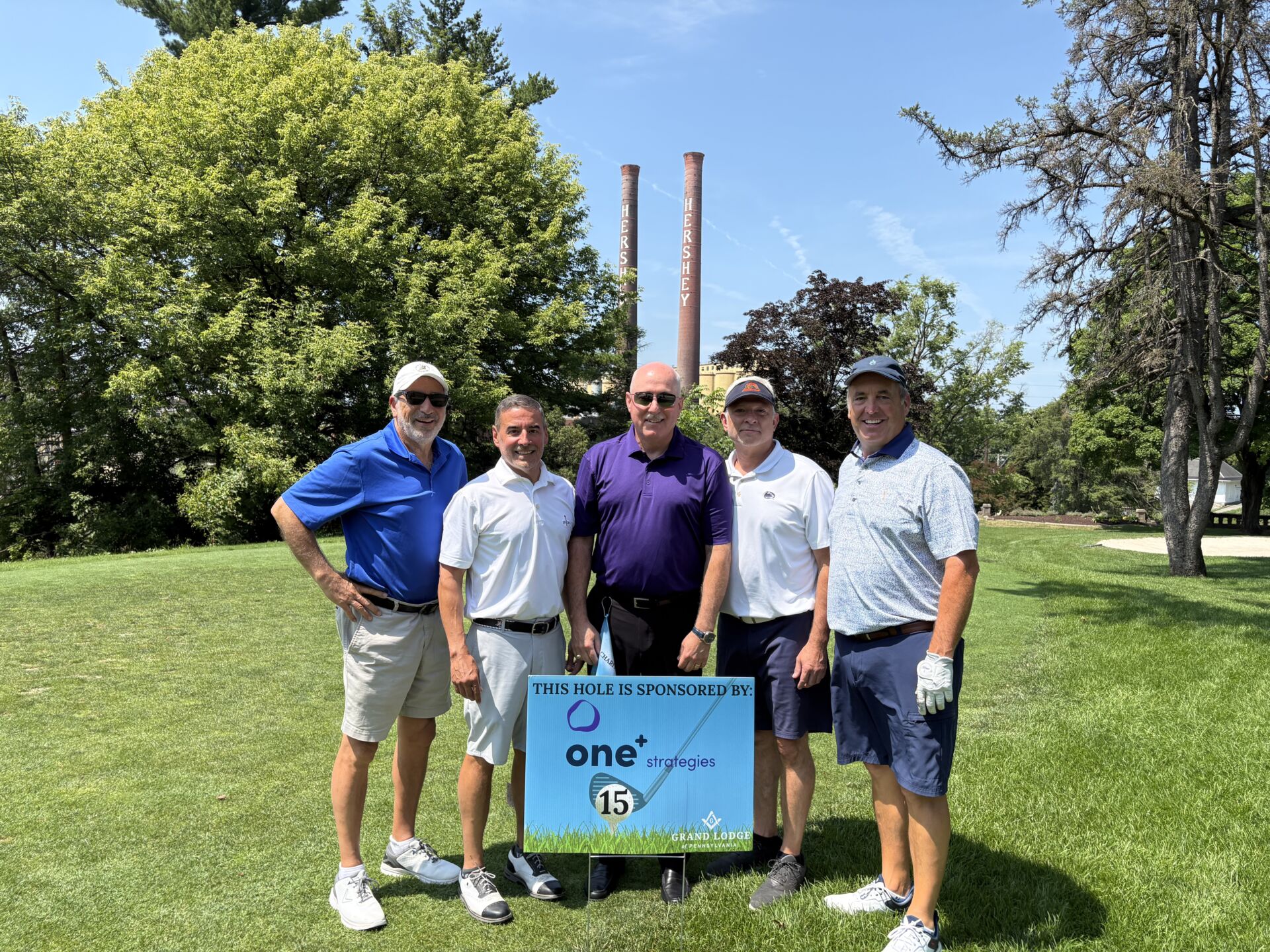 Group of Masons dressed in golfing attire with Grand Master Brink standing in the center. A sign reading "This hole is sponsored by One+ Strategies is in front of them.